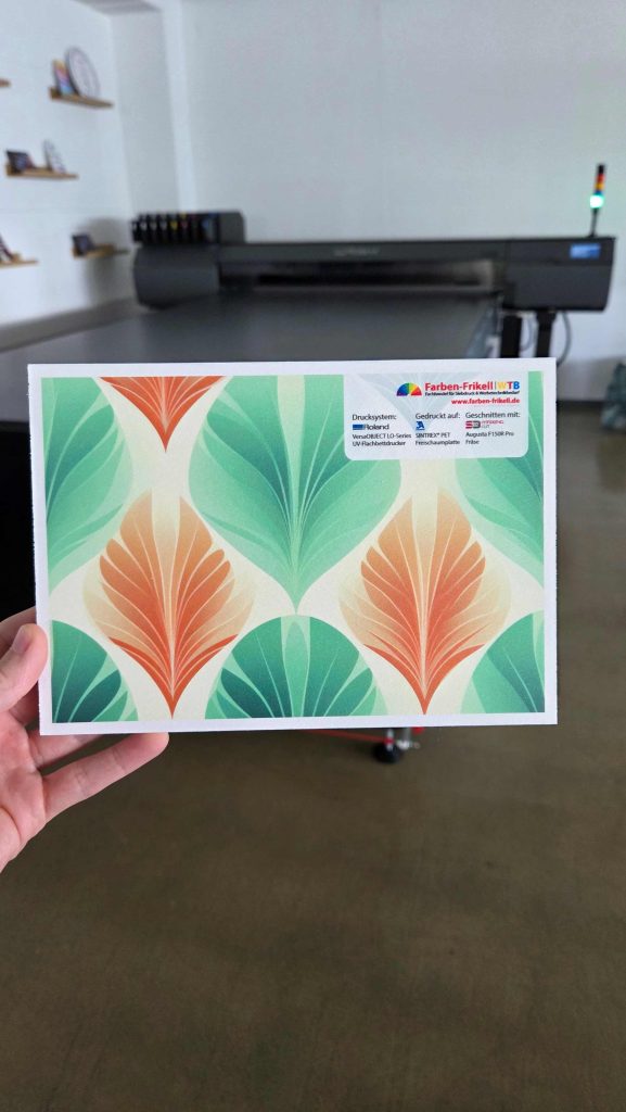 PET-Hartschaumplatte "SINTREX" bedruckt mit einem floralem Bild. Der UV-Flachbettdrucker LO-640 von Roland DG ist im Hintergrund. Die Platte hat saubere Kanten und wurde mit der Fräs- und Schneidmaschine F150R Pro gefräst. Die Musterplatte von Farben-Frikell | WTB wird in der Hand gehalten und hat oben rechts ein Label mit Benennung der verwendeten Maschinen.