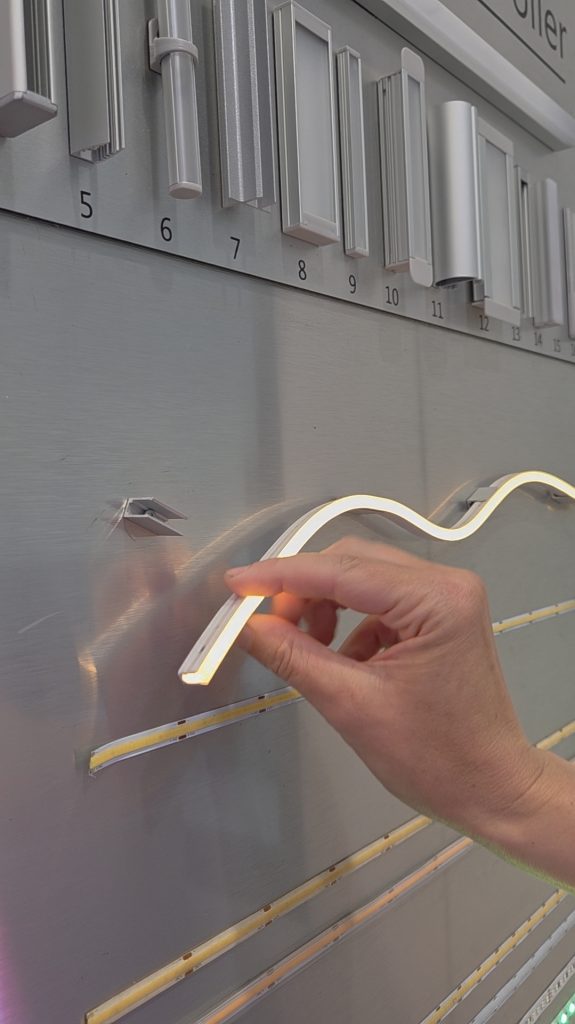 Aluminiumhalter für LED Flex Neon von Farben-Frikell | WTB. Eine Hand hält die LED Flex Neon, die wellig an einer Aluminiumplatte befestigt ist. Das letzte Ende ist noch nicht im Aluminiumhalter eingelegt.
